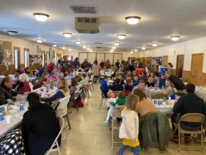 A crowded church fellowship hall with people sitting at long dining tables.