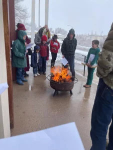 Children and adults gathered around a burning fire pit outside during winter.